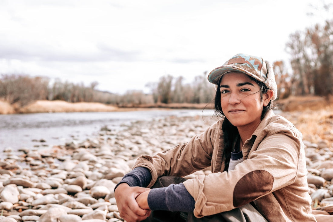 Person sitting on a rocky riverbank wearing outdoor clothing and a cap, enjoying a calm moment in nature.
